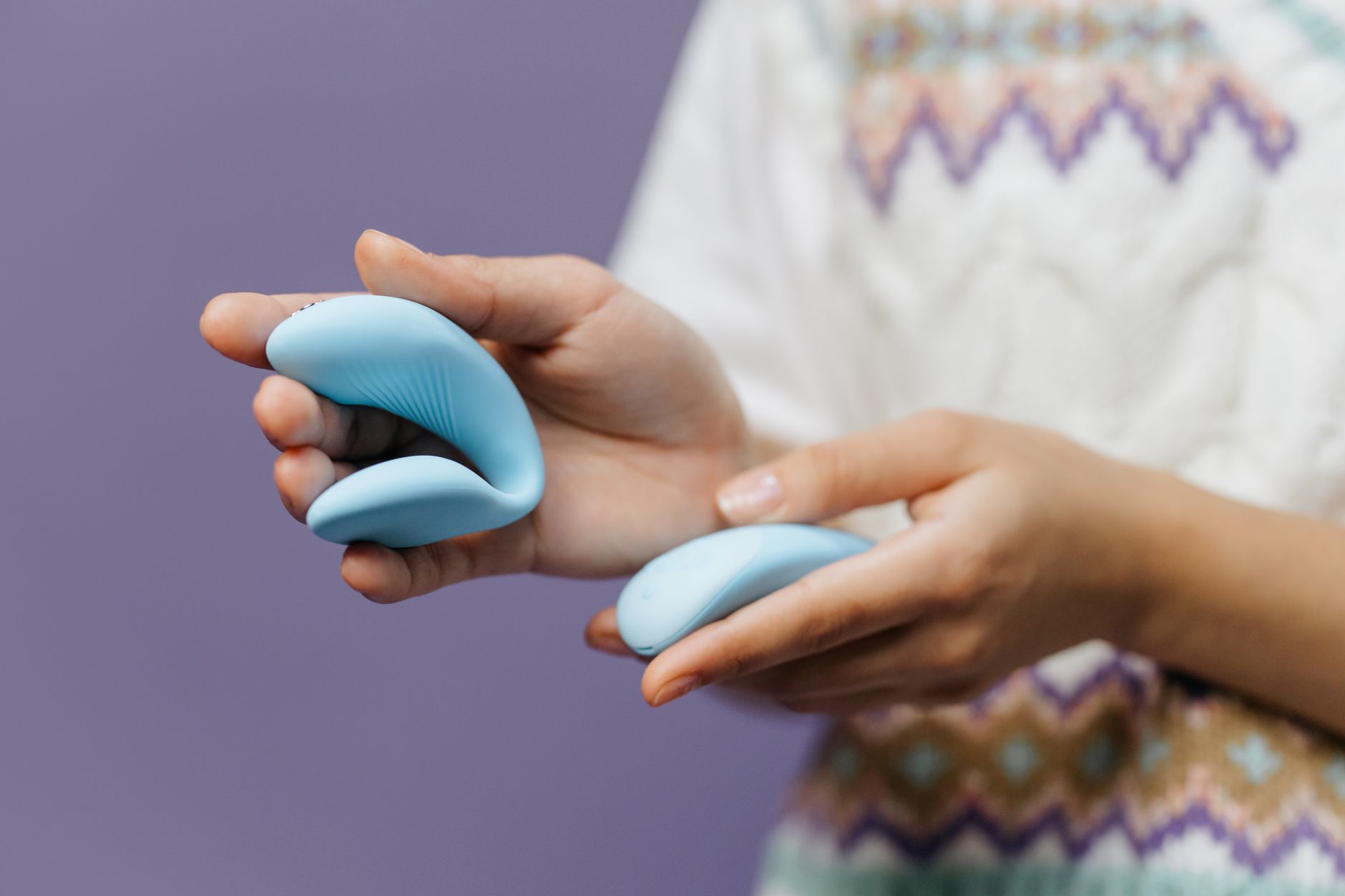 Close-up hands holding a sleek blue clitoral vibrator against purple background, emphasizing intimate self-care.