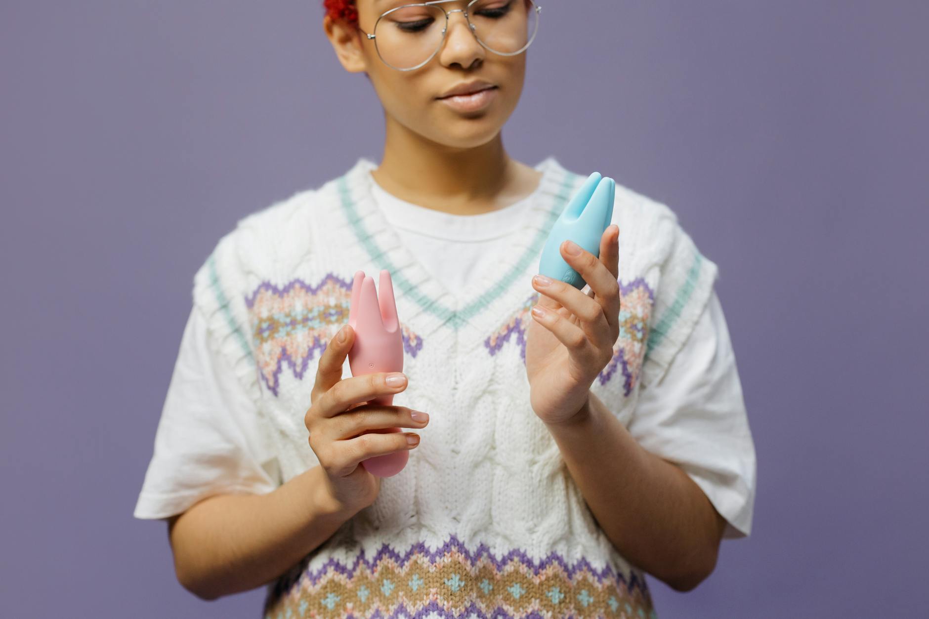 Woman holding blue and pink silicone vibrators thoughtfully against soft background